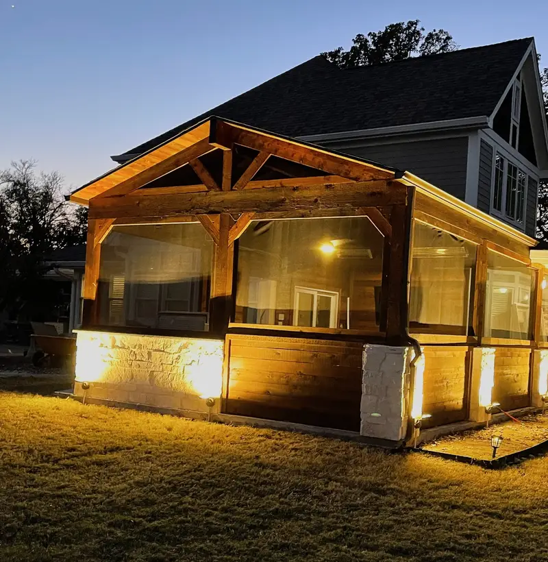 Screened cedar porch with a timber gable and white stone base, glowing under landscape lights at dusk