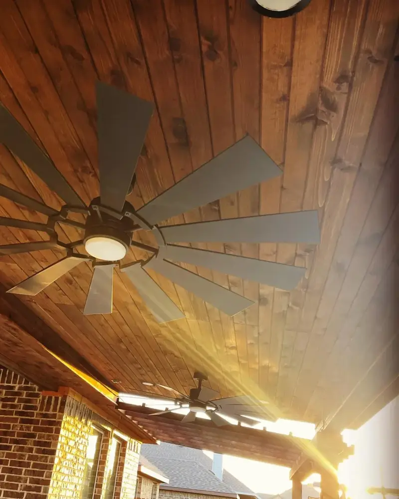 Underside of a stained cedar tongue-and-groove patio ceiling with a large windmill-style fan, a brick wall below and low sun flaring past the posts