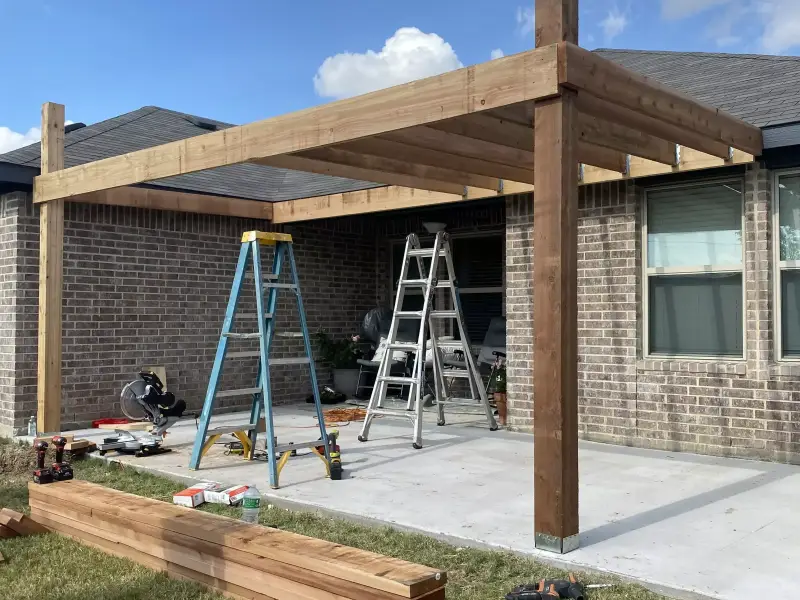 Cedar pergola framing going up against a brick home: posts and beams set on a concrete slab, two ladders and a miter saw underneath, fresh cedar boards stacked on the lawn
