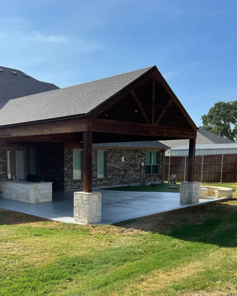 Gable patio cover with dark-stained wood beams and stone-clad column bases over a gray concrete patio, with a stone outdoor kitchen counter and a curved stone seat wall, attached to the back of a brick home on a sunny day
