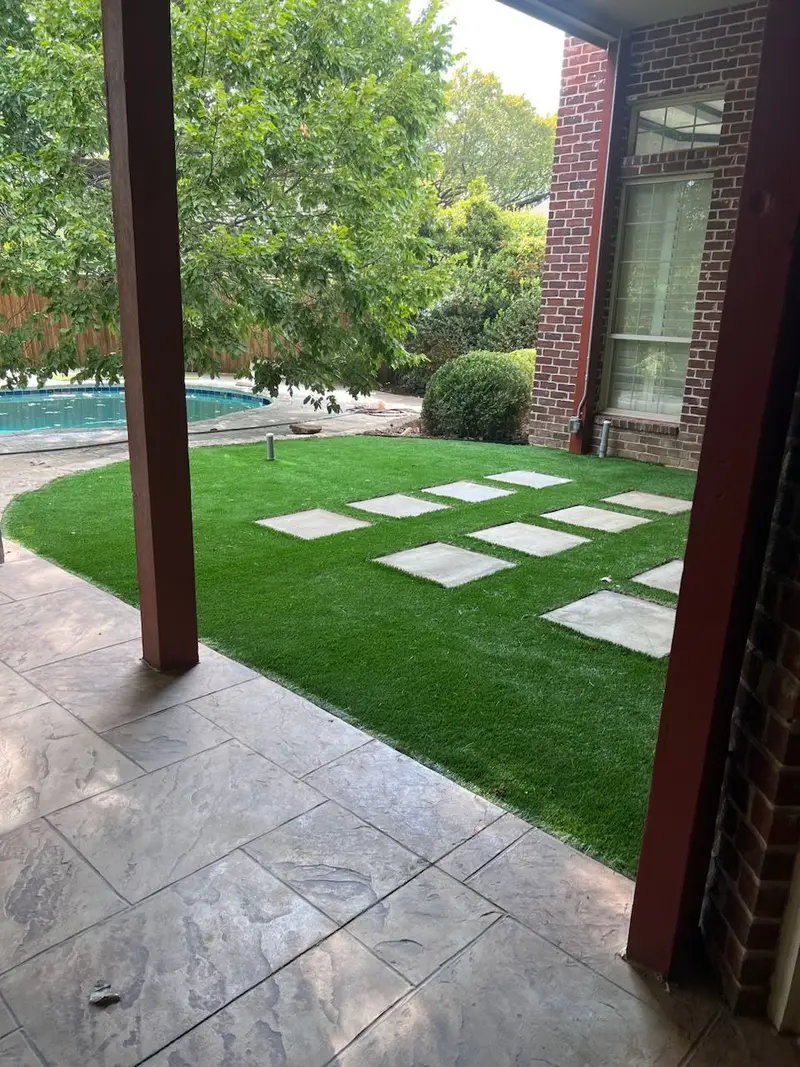 Artificial turf with square stepping pads beside a stamped-concrete patio and a swimming pool.