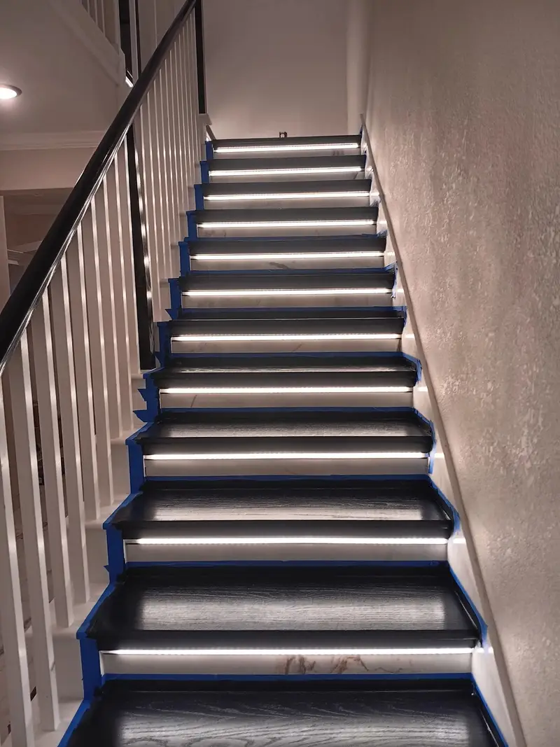 Interior staircase renovation in progress with dark treads, white balusters, a black handrail, LED step lighting and blue masking tape.
