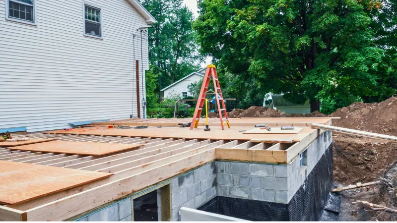 Floor joists and subfloor under construction on a block foundation beside an existing house.