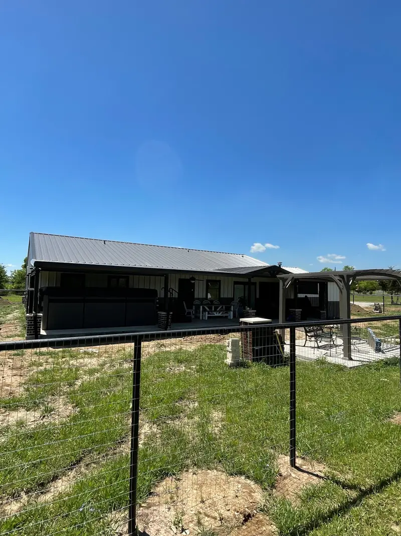 Dark barndominium-style home with a metal roof and covered porch behind a wire fence.