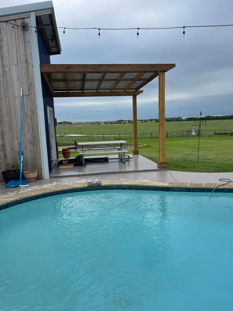 Swimming pool beside a timber pergola and a board-and-batten building, with open lawn beyond.