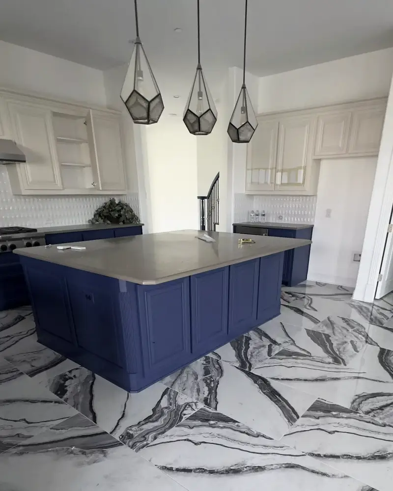 Kitchen with a navy-blue island under three geometric glass pendant lights, cream cabinets, and glossy black-and-white 2 by 4 ft marble floor tiles running through the room