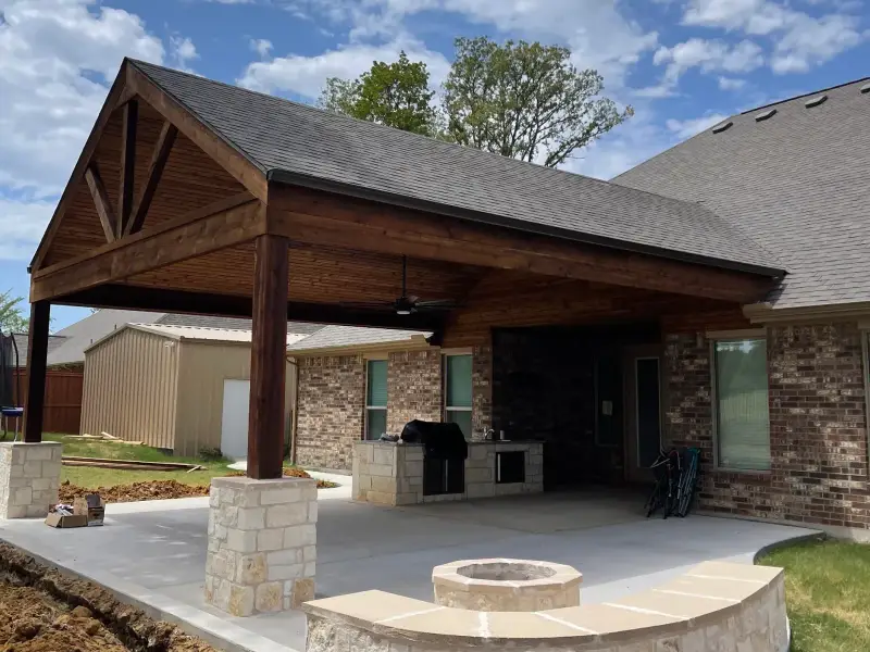 Gable patio cover with exposed wood beams and stone-clad columns over a new concrete patio, a stone outdoor kitchen with a grill under it, and a round stone fire pit inside a curved stone seat wall; brick home, blue sky, freshly graded yard