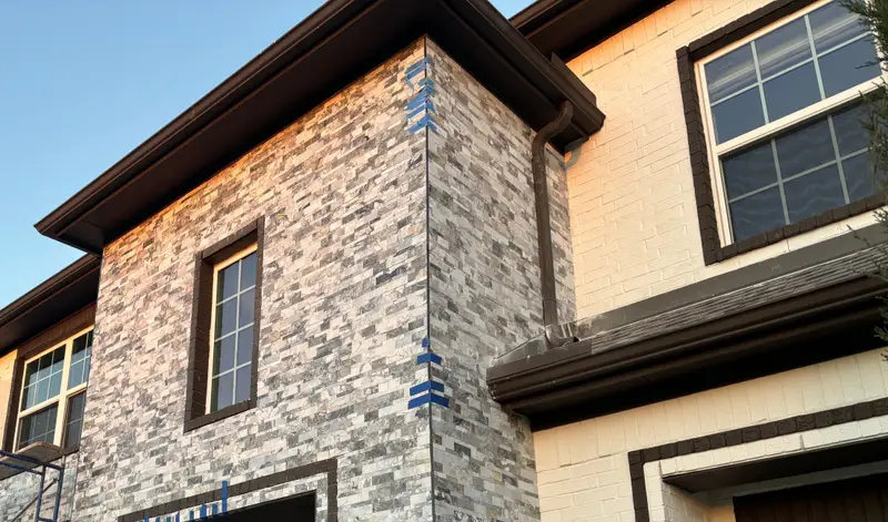 Two-story home getting a stacked-stone facade in low golden sunlight: gray and white split-face stone wraps a square tower with a tall dark-framed window, blue layout tape still on the corner, bronze trim and gutters, a painted brick wing at right and scaffolding at the far left