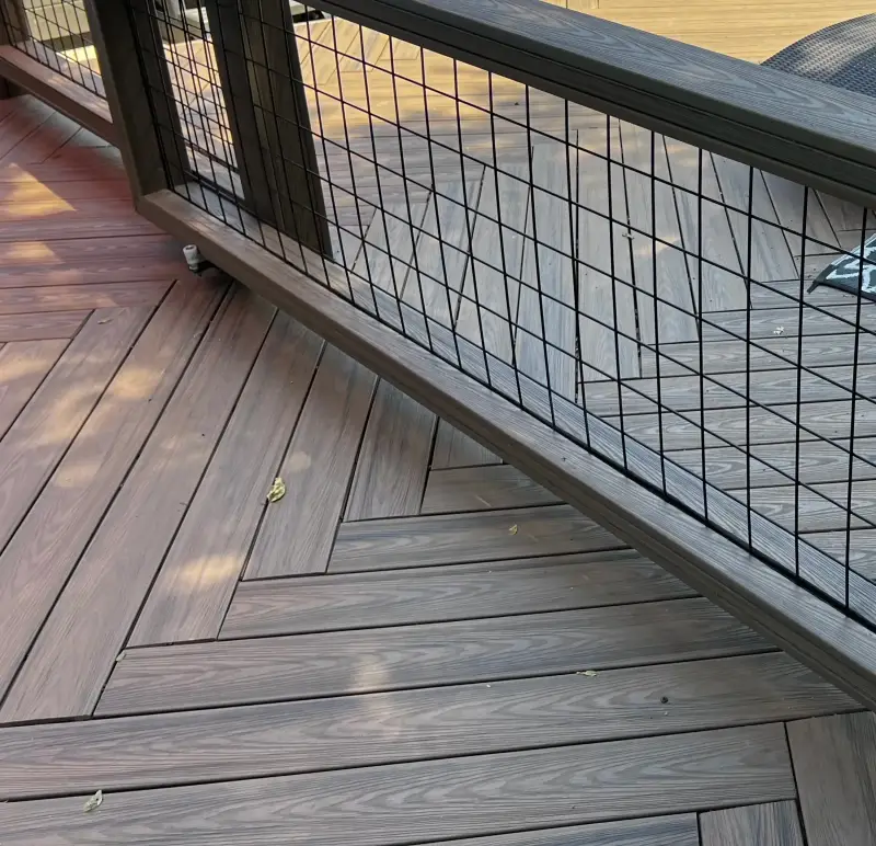 Close view of brown composite deck boards laid in a herringbone pattern, edged by a dark railing with black hog-wire panels