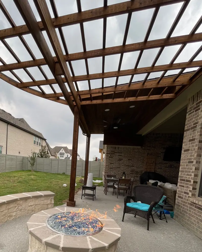 Stained wood pergola over a patio with a round stone fire pit burning with blue fire glass, a curved stone seat wall and outdoor seating