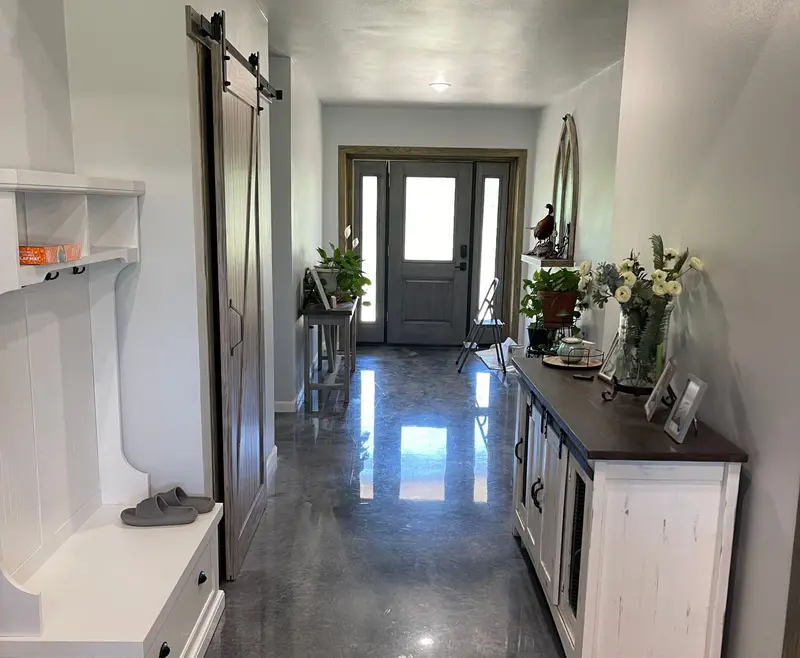 Long hallway with a glossy gray epoxy floor reflecting the light from the front door, a sliding barn door, a white mudroom bench and a white console table with flowers