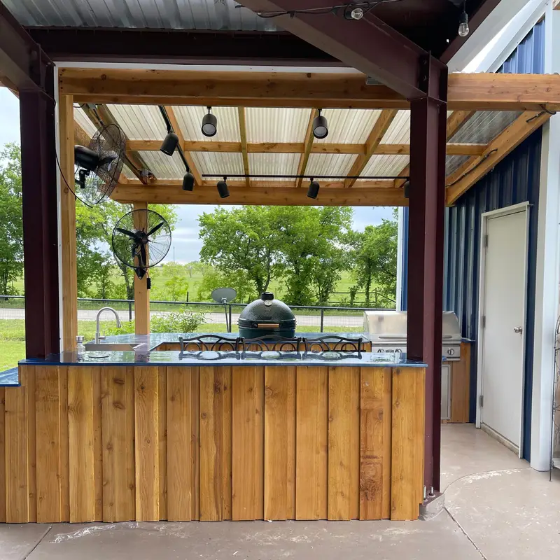 Outdoor kitchen pavilion with a cedar-clad bar, grill and kamado smoker under a metal roof, looking out over open pasture