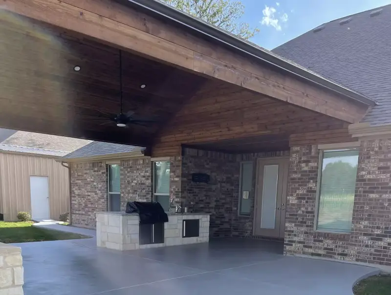 Covered patio with a stained wood ceiling and gray coated floor, and a stone outdoor kitchen with a grill against the brick house