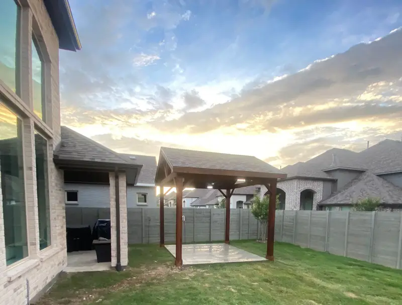 Backyard patio pavilion with stained wood posts on a new concrete pad, fresh lawn and a gray fence beside a light brick two-story home, under a sunset sky