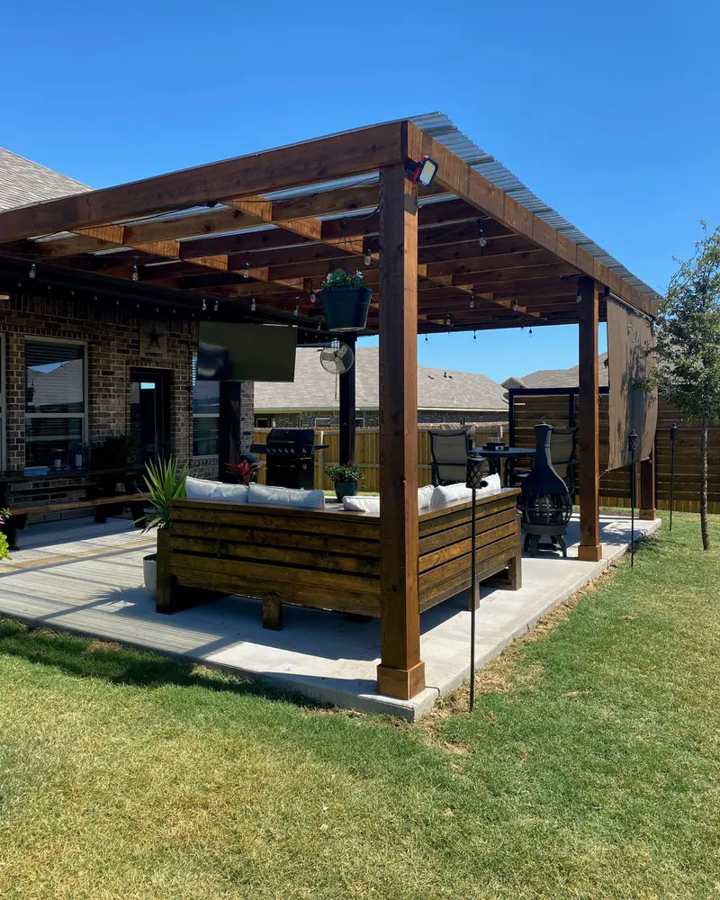 Stained cedar pergola patio cover on a brick home, with a built-in wood sofa and a dining set on the concrete slab, green lawn and clear blue sky