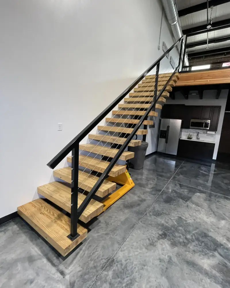 Floating wood stairs with a black steel rail rising over a glossy gray metallic epoxy floor, a kitchenette under the loft